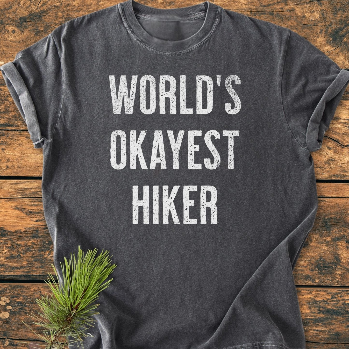 Heather-gray World's Okayest Hiker Vintage Wash Tee with bold white text on rustic wood background, featuring pine sprig accent. Made from soft-washed, garment-dyed 100% ring-spun cotton for comfort and durability.