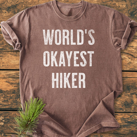 World's Okayest Hiker Vintage Wash Tee, brown, short-sleeved with distressed white lettering, laid flat on rustic wood. Pine needles beside it emphasize an outdoor theme.