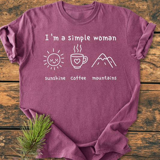I'm A Simple Woman Vintage Wash Tee on wooden surface, featuring white line art of a sun, coffee mug, and mountains. Includes a pine sprig for styling. Soft, relaxed fit, 100% cotton.