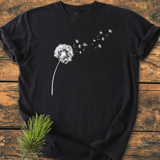 The Dandelion Vintage Wash Tee features a black crew-neck with a white dandelion graphic, sleeves casually rolled, set on a wooden surface, accentuated by a pine sprig.