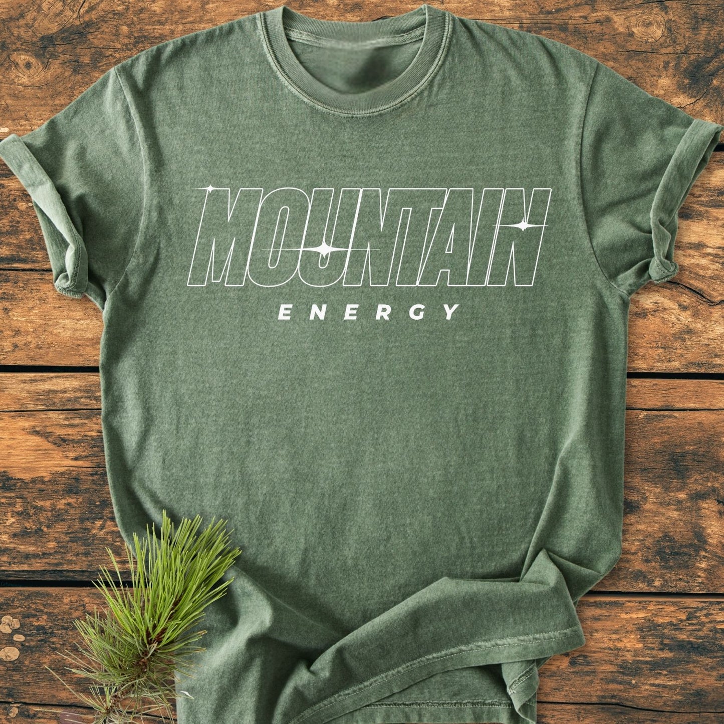 Mountain Energy Vintage Wash Tee in heather-green, featuring bold MOUNTAIN and ENERGY lettering, laid flat with a pine twig accent, highlighting its rustic and outdoorsy appeal on a wooden surface.