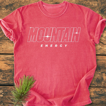 Mountain Energy Vintage Wash Tee in washed-out red, screen-printed with MOUNTAIN ENERGY text. Laid flat on a wooden surface, enhanced by a pine branch accent, showcasing its relaxed, soft-washed fit.