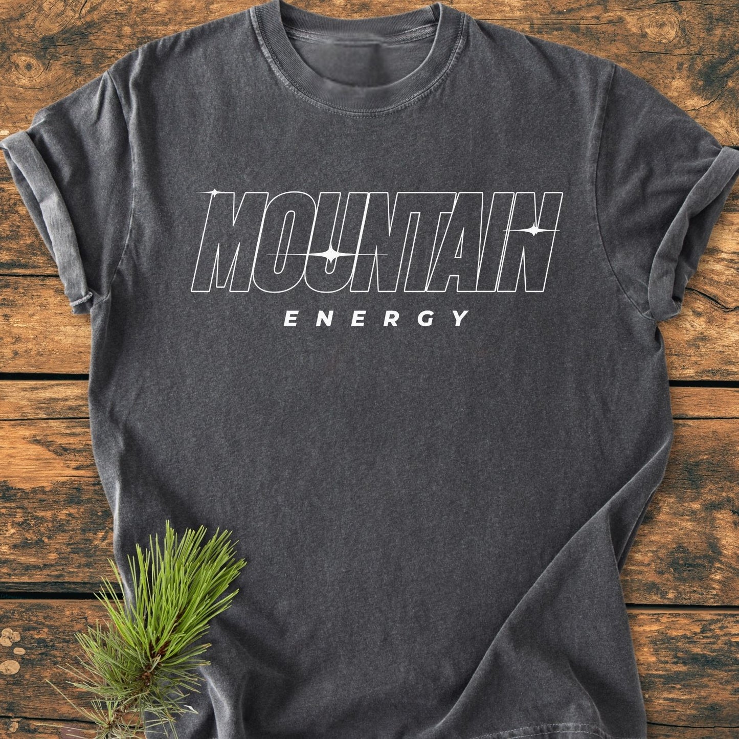 Mountain Energy Vintage Wash Tee in charcoal-gray, featuring MOUNTAIN ENERGY text, displayed on wood surface with pine branch. Soft-washed, relaxed fit, durable with double-needle stitching, made from ring-spun cotton.
