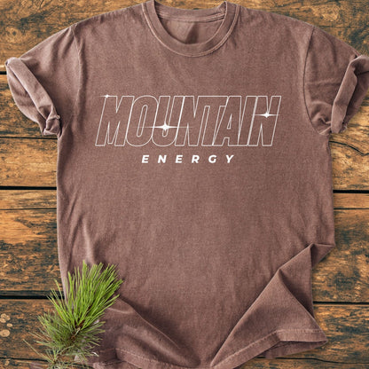 Mountain Energy Vintage Wash Tee features a brown, short-sleeved design with MOUNTAIN ENERGY graphic, laid on a wooden surface with a pine sprig, showcasing its relaxed, durable style.