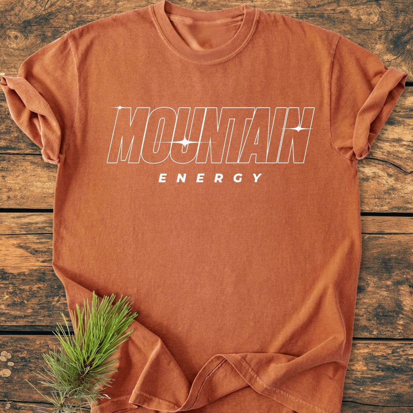 Mountain Energy Vintage Wash Tee: Rust-orange cotton tee with 'MOUNTAIN ENERGY' in white outline font, star-sparkle accents, rolled sleeves, on wooden surface, pine sprig under hem.