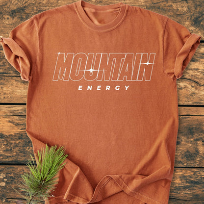 Mountain Energy Vintage Wash Tee: Rust-orange cotton tee with 'MOUNTAIN ENERGY' in white outline font, star-sparkle accents, rolled sleeves, on wooden surface, pine sprig under hem.