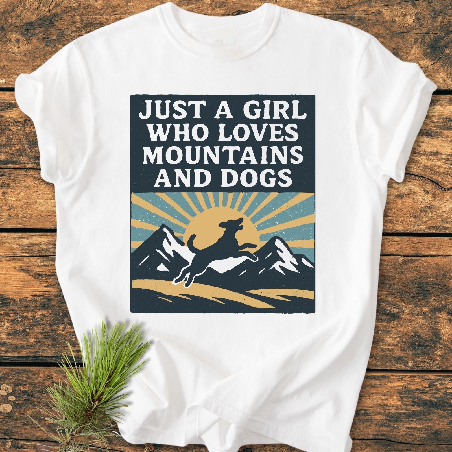 Just A Girl Who Loves Dogs - Vintage Wash Tee featuring a graphic of mountains, a leaping dog, and a sunrise, laid on wood with a pine sprig nearby.