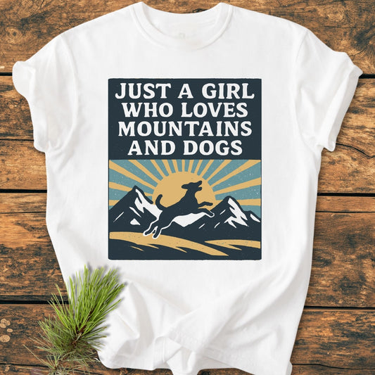 Just A Girl Who Loves Dogs - Vintage Wash Tee featuring a graphic of mountains, a leaping dog, and a sunrise, laid on wood with a pine sprig nearby.