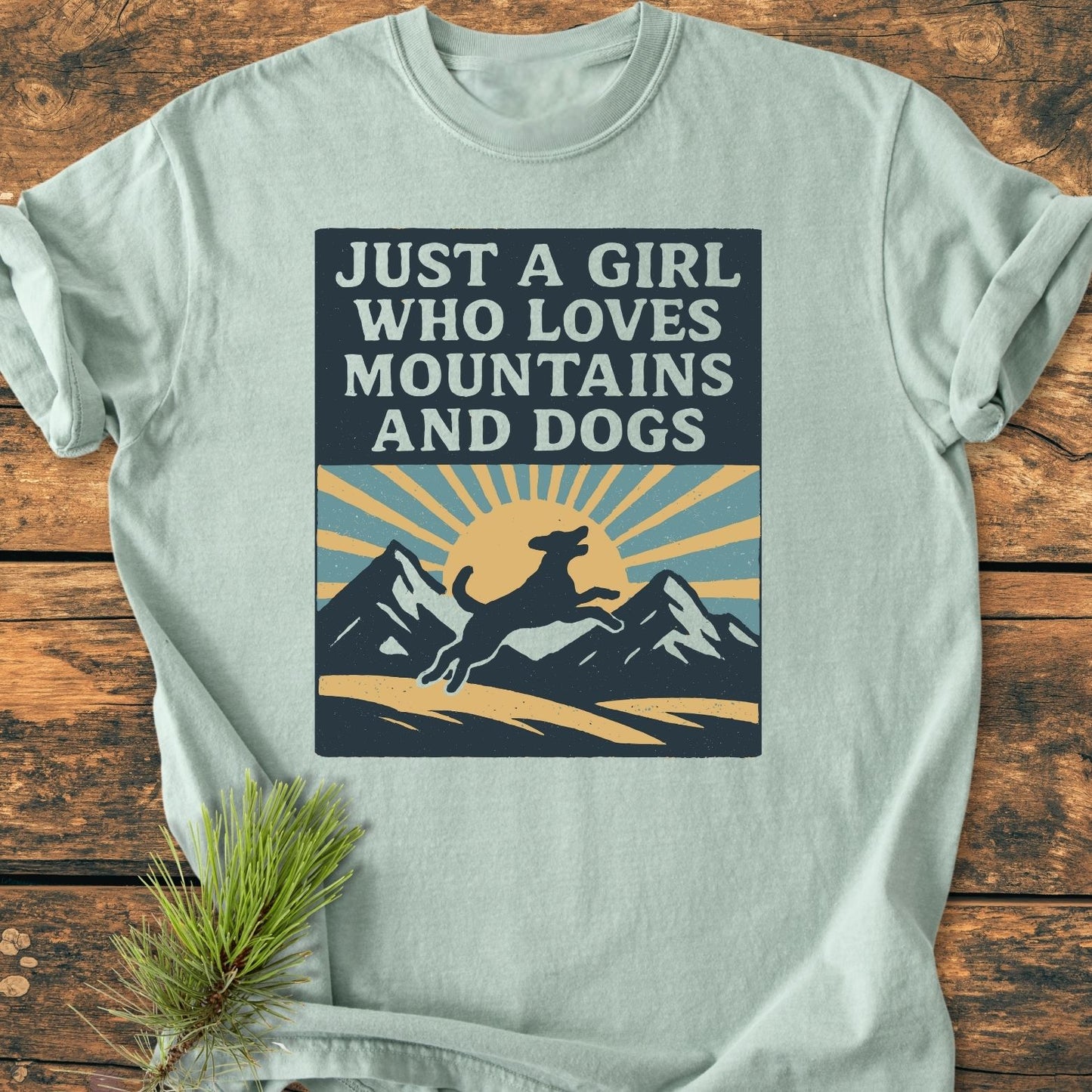 Just A Girl Who Loves Dogs - Vintage Wash Tee; light-teal T-shirt featuring a retro dog and mountain print, laid on a wooden surface with a pine sprig accent.