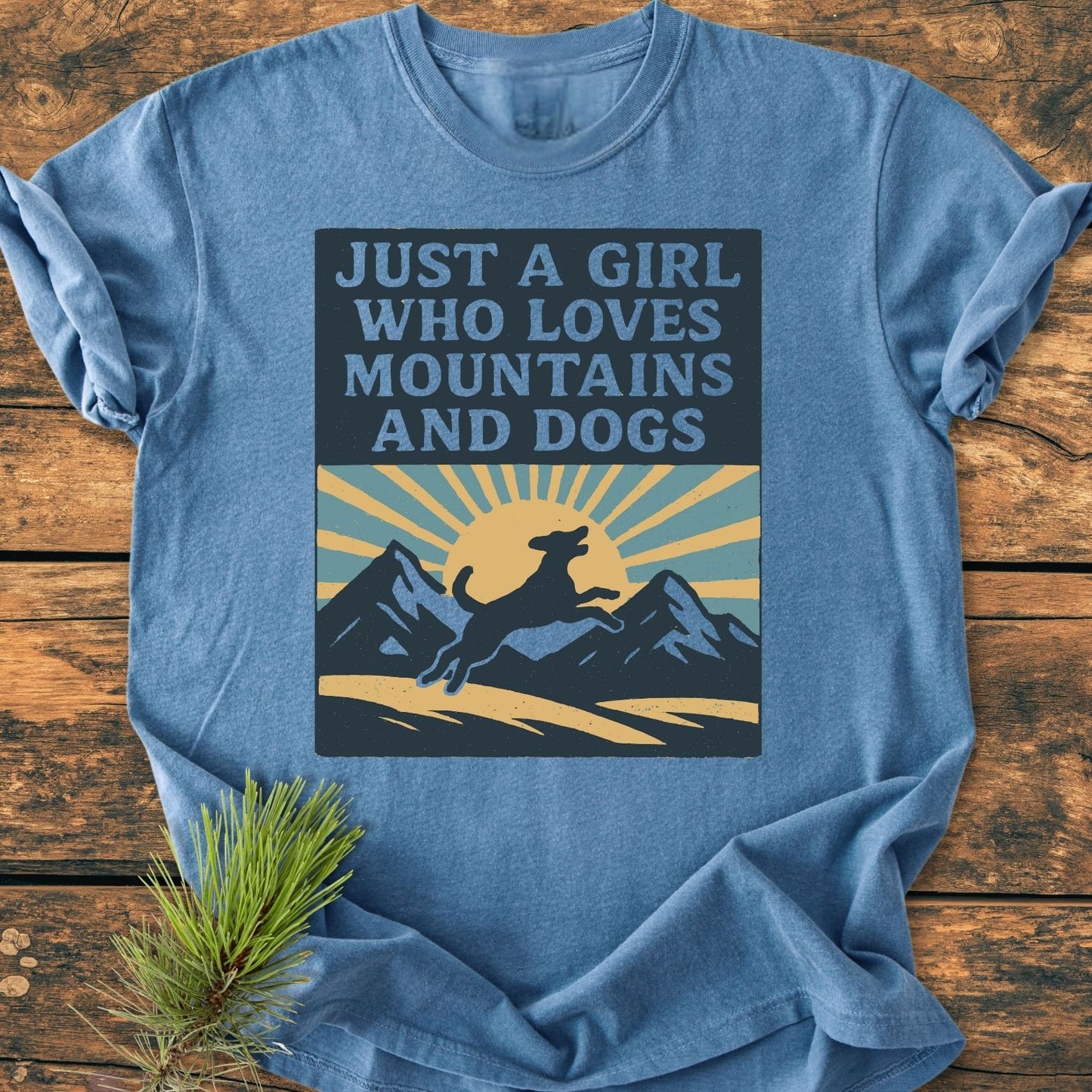 Just A Girl Who Loves Dogs - Vintage Wash Tee featuring a mountain graphic and leaping dog on a heather-blue T-shirt, laid on a wooden surface with pine sprig, showcasing a cozy, relaxed fit.