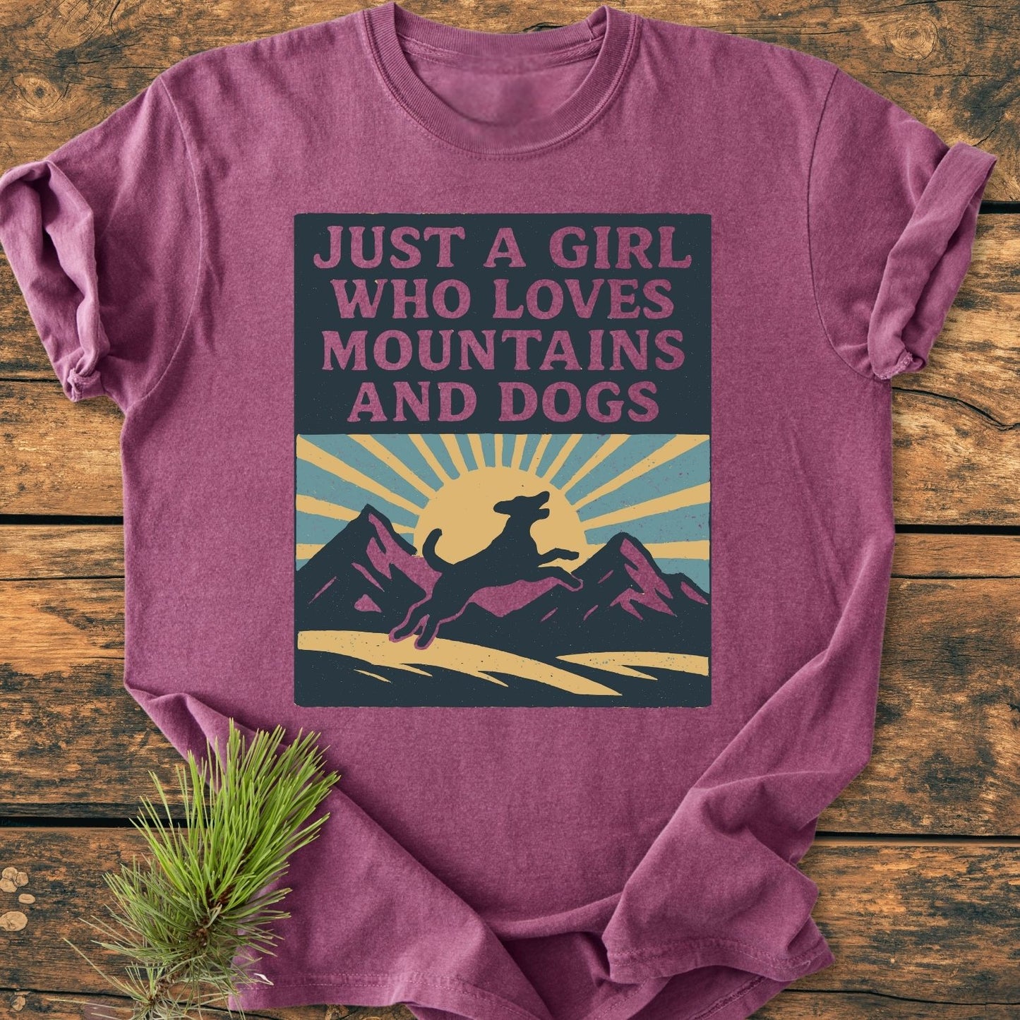 Just A Girl Who Loves Dogs - Vintage Wash Tee features a retro graphic of mountains and a leaping dog, laid on a wooden surface with a pine branch accent.