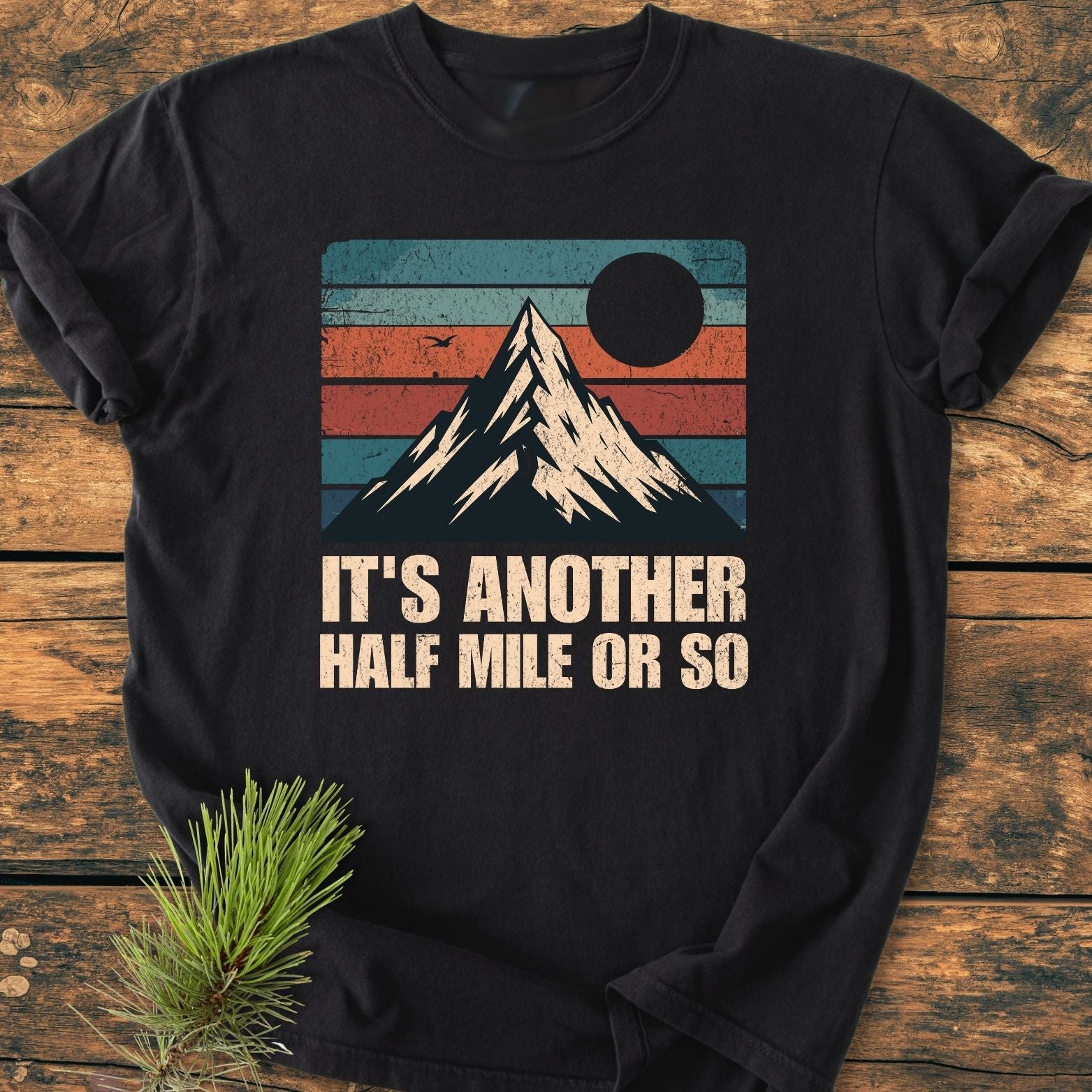 The It's Another Half Mile - Vintage Wash Tee features a snow-capped mountain graphic on a black T-shirt, set against a retro-striped background, with a distressed slogan below, laid on a wooden surface.