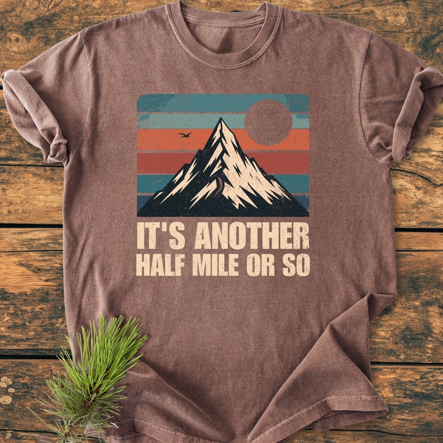 It's Another Half Mile - Vintage Wash Tee features a stylized mountain over retro color bands, with text below. Laid on wood with pine sprig, emphasizes comfort and durability.