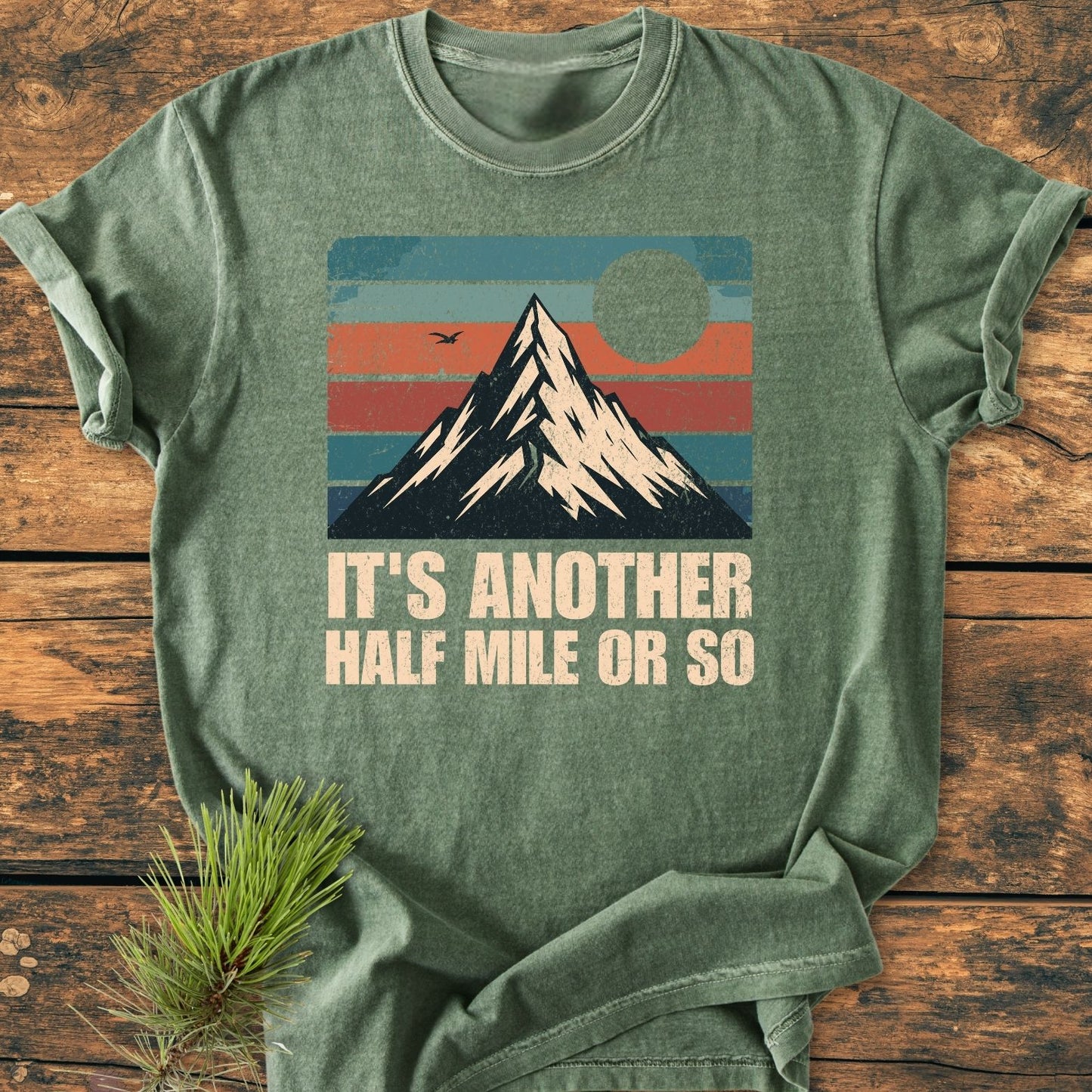 It's Another Half Mile - Vintage Wash Tee lays on wood, featuring a snowy mountain graphic with teal, orange stripes, sun, bird, and slogan. Pine sprig accentuates the cozy, garment-dyed shirt.