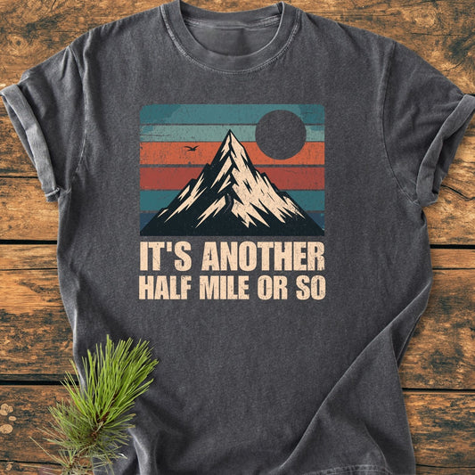 Dark-gray 'It's Another Half Mile - Vintage Wash Tee' with retro mountain graphic, featuring teal, orange, red, and blue stripes, laid flat on wood. Text below reads, 'IT’S ANOTHER HALF MILE OR SO.'