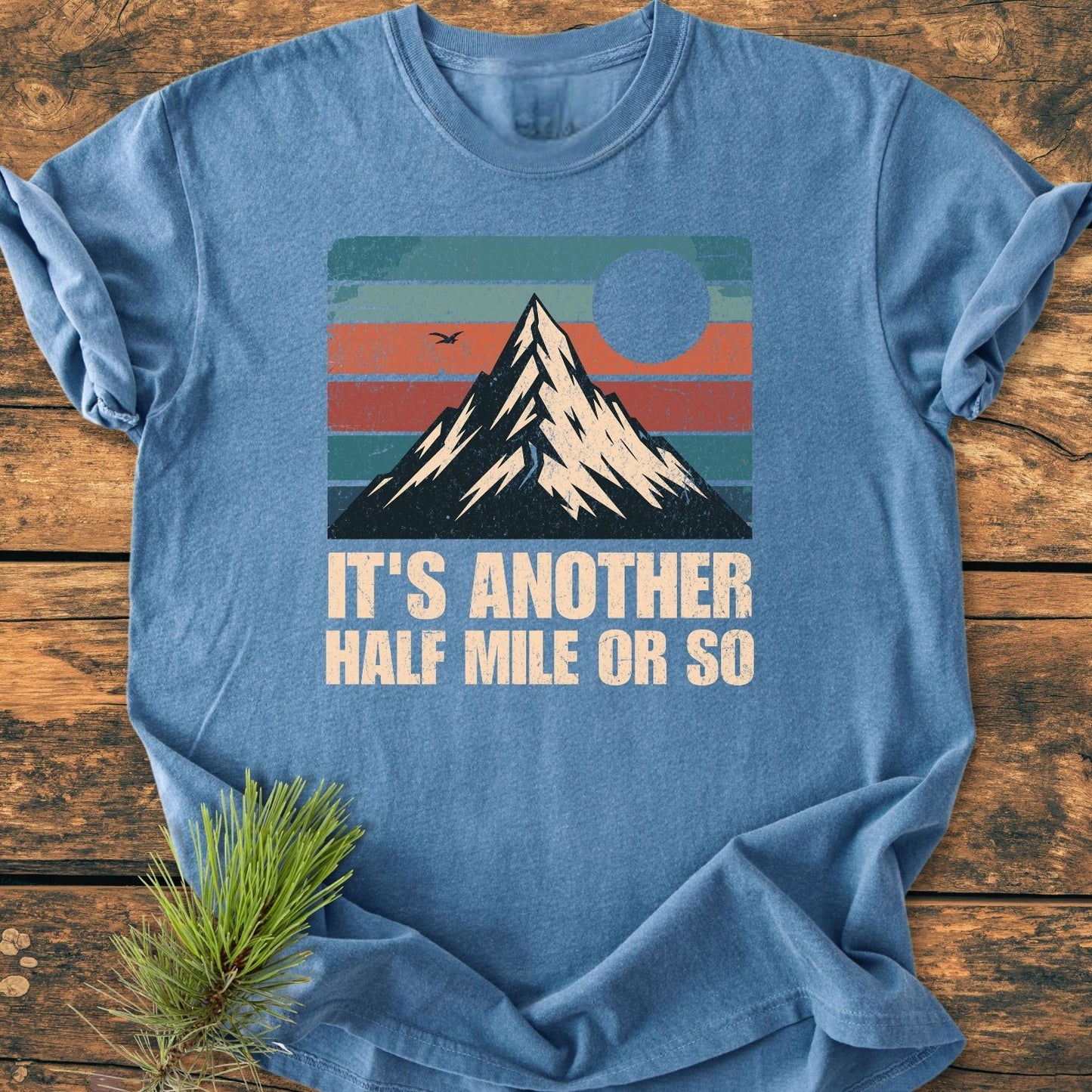 The It's Another Half Mile - Vintage Wash Tee features a retro mountain graphic with a sun/moon, set against muted stripes on a heather-blue background, conveying an outdoorsy vibe.