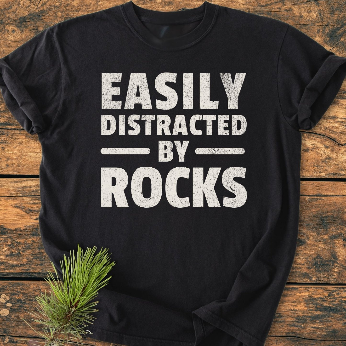 Easily Distracted By Rocks - Vintage Wash Tee on rustic wood, featuring large distressed white text. Styled with pine needles, showcasing the soft-washed, relaxed fit and durable double-needle stitching.