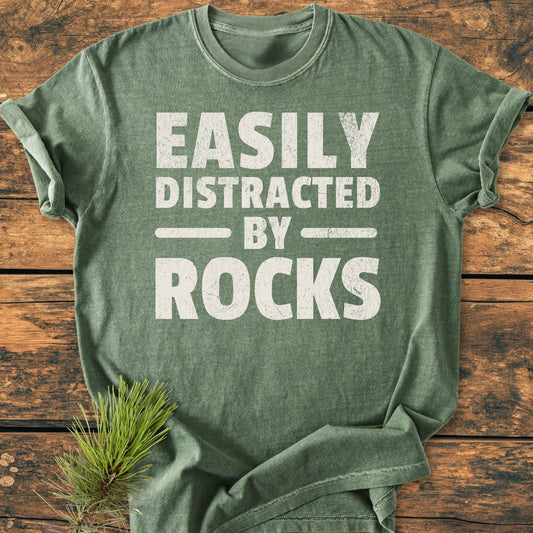 Easily Distracted By Rocks - Vintage Wash Tee, heather-green crew-neck with distressed white print, laid on rustic wood, featuring pine sprig accent. Soft, relaxed-fit, 100% ring-spun cotton, durable stitching.