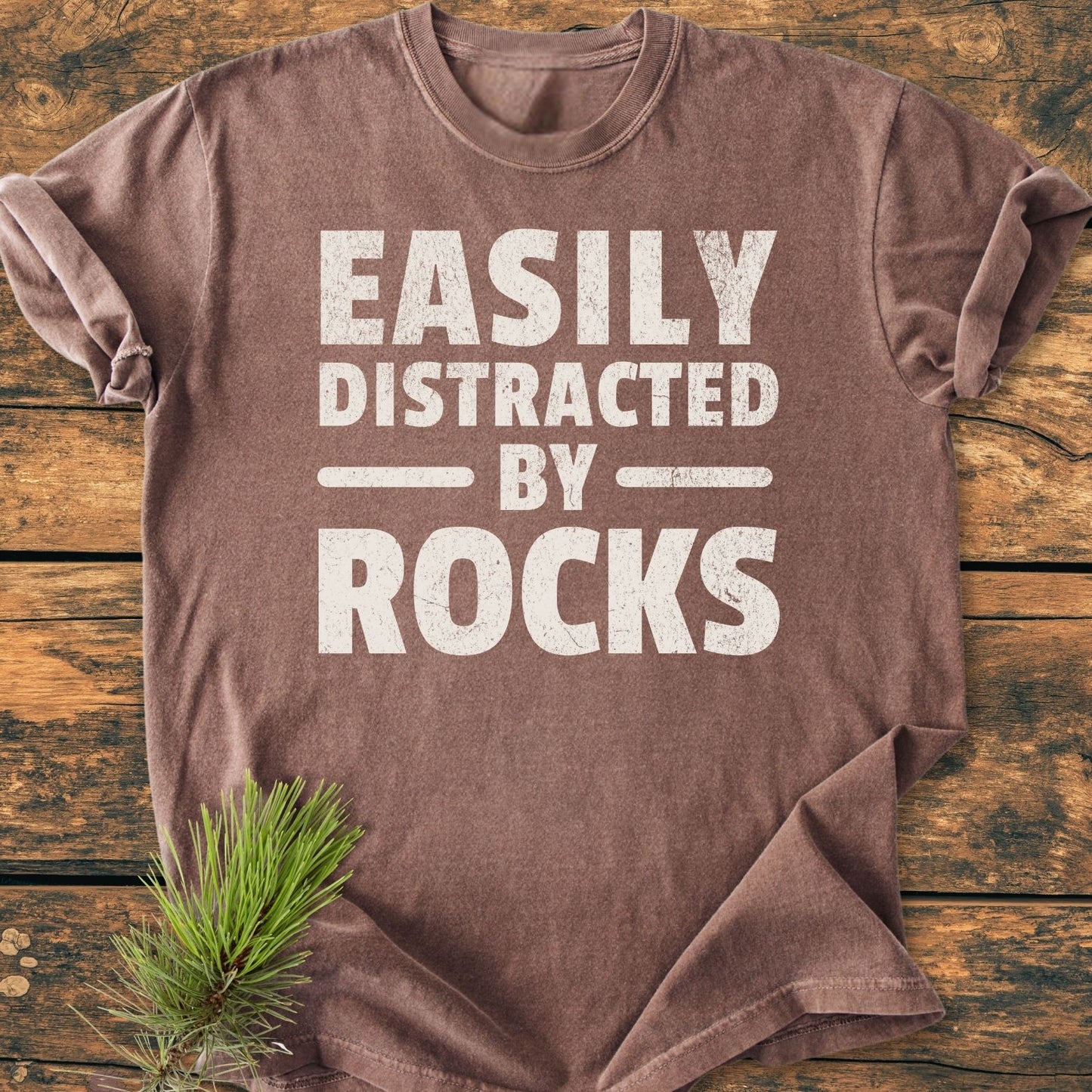 Easily Distracted By Rocks - Vintage Wash Tee displayed on wood surface, featuring distressed white text. Soft, relaxed fit, short-sleeved crew-neck, styled with pine needles accent for a rustic touch.