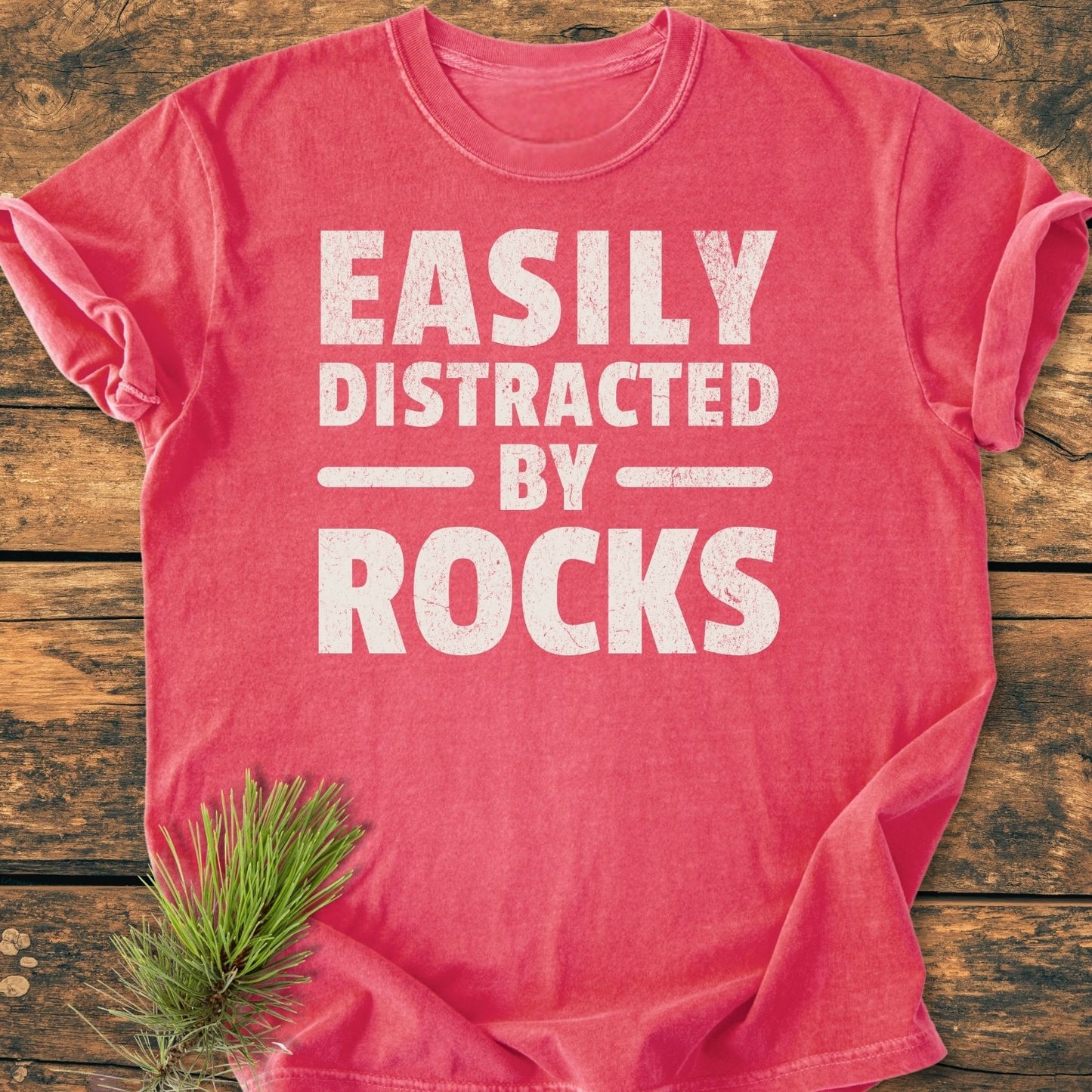 Easily Distracted By Rocks - Vintage Wash Tee flat-lay, displaying distressed white text on a soft-washed red surface, with pine needles accentuating the garment's rustic charm on a wooden background.