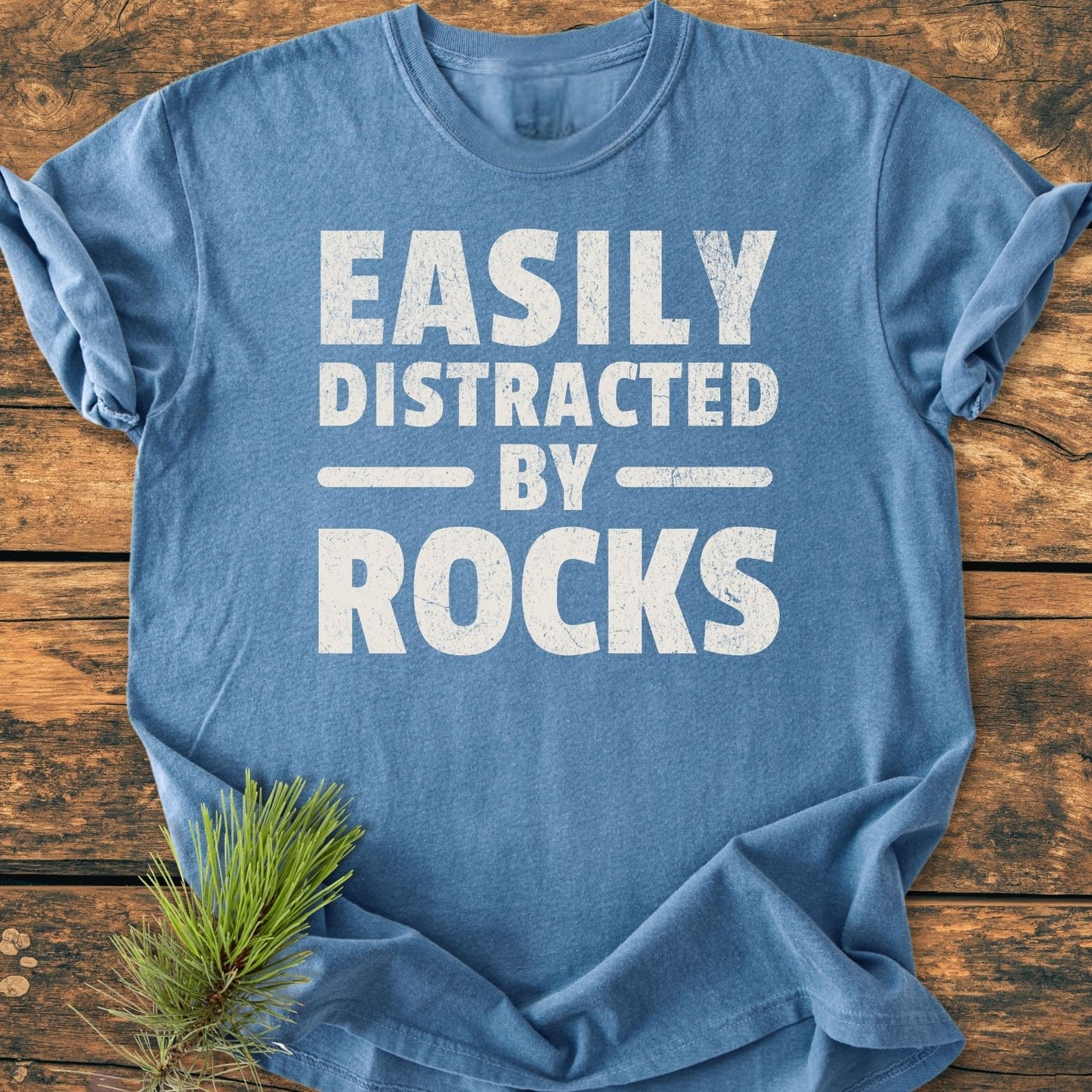 The Easily Distracted By Rocks - Vintage Wash Tee features a heather-blue color with rolled sleeves, printed white text, and a pine sprig accent on a rustic wooden surface.