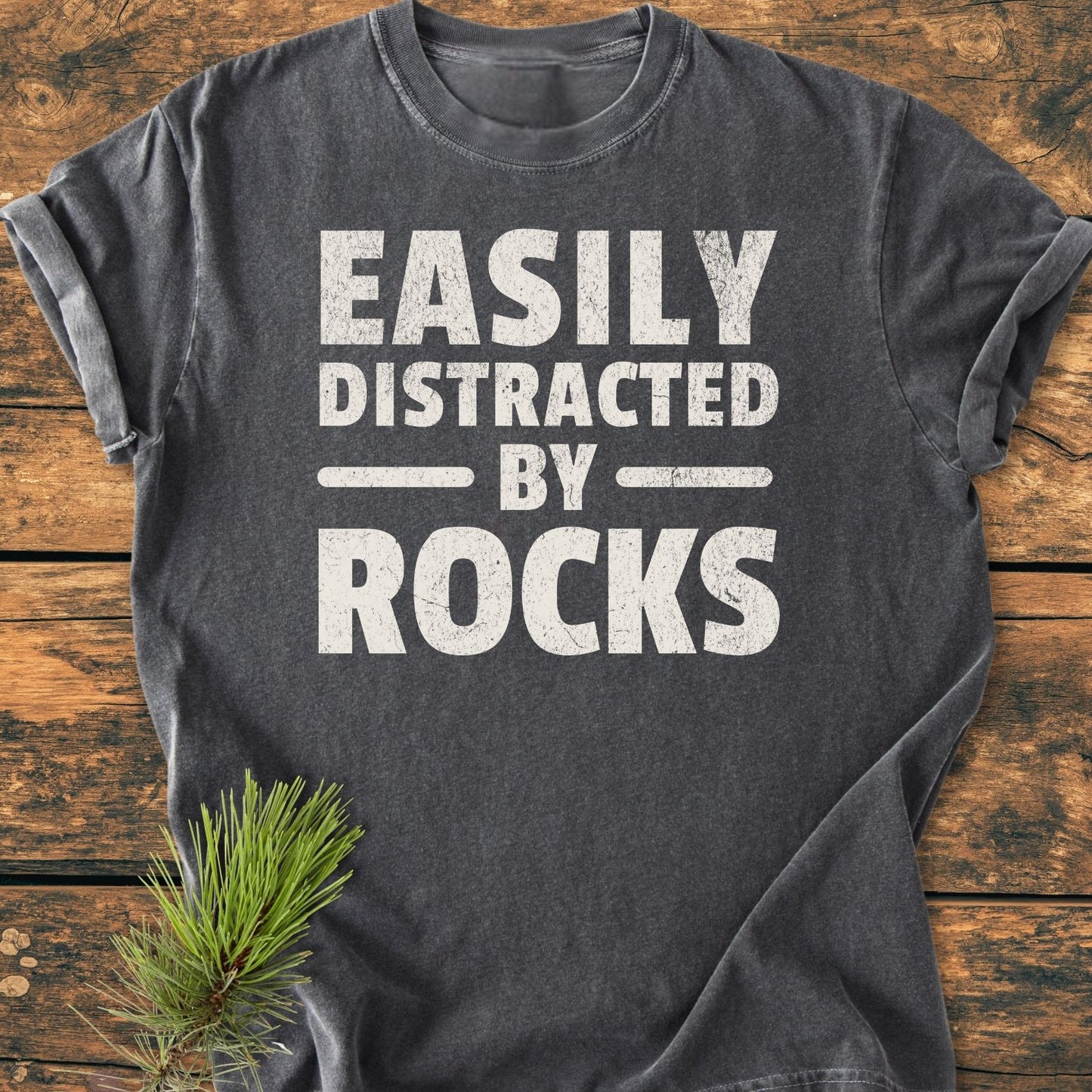 Easily Distracted By Rocks - Vintage Wash Tee on wooden surface, sleeves rolled up, bold distressed white text. A small pine branch accents the scene, highlighting the tee's relaxed, durable design.