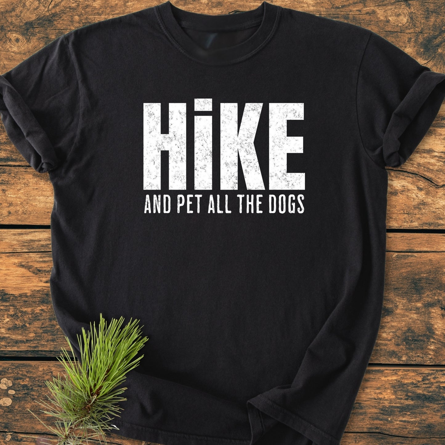 Hike And Pet All The Dogs - Vintage Wash Tee: Black T-shirt with a white slogan, laid on wood, featuring distressed lettering. A pine sprig accents the soft, relaxed-fit, garment-dyed cotton shirt.