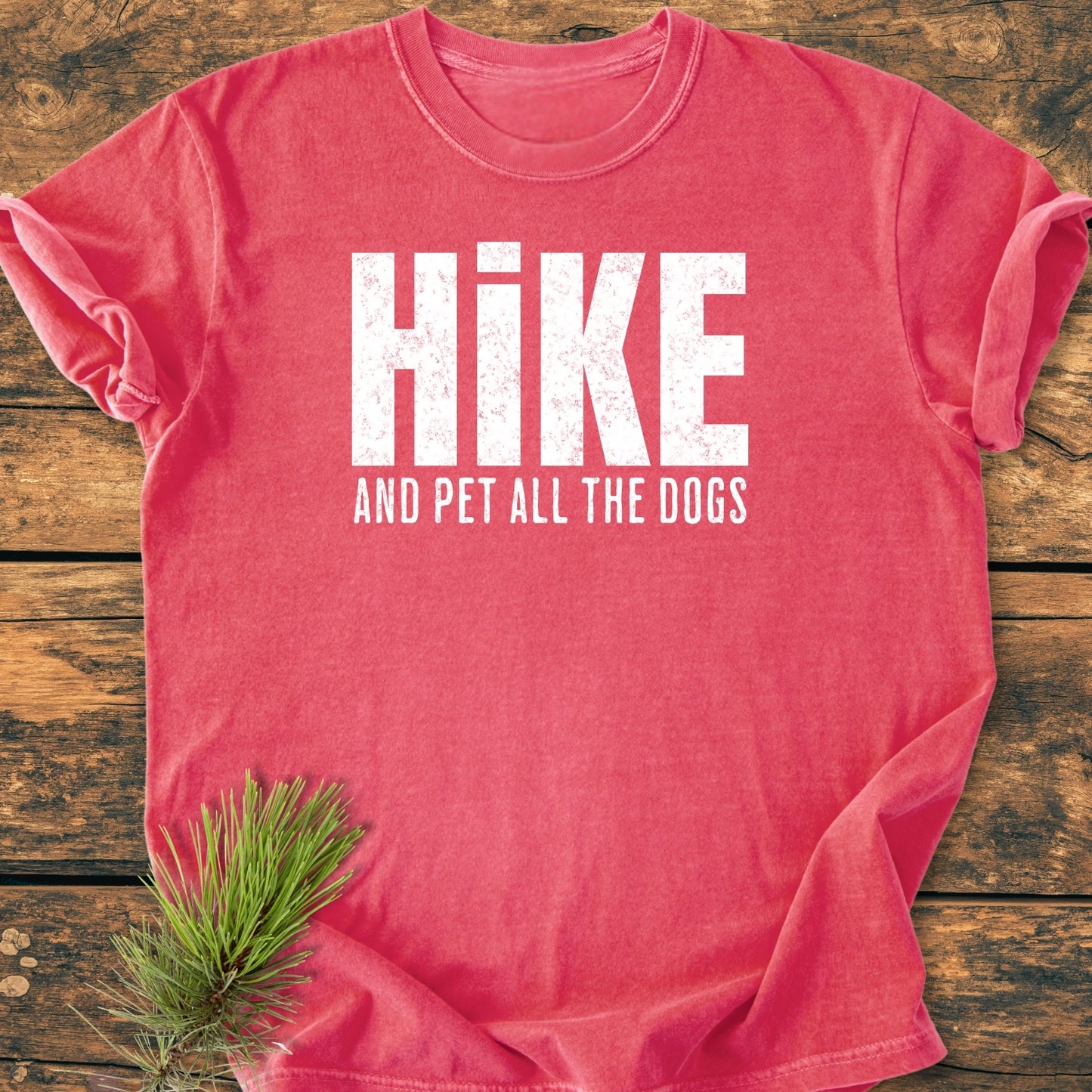 Hike And Pet All The Dogs - Vintage Wash Tee in heather-red lays flat on wood, showcasing the distressed white text and a pine needle accent for a woodland touch.