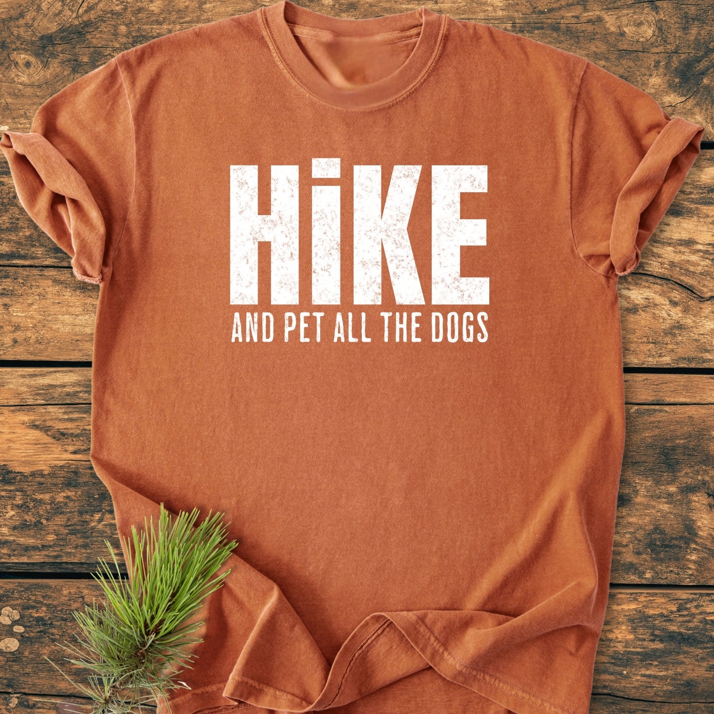 Hike And Pet All The Dogs - Vintage Wash Tee: Rust-orange with HIKE and AND PET ALL THE DOGS in white. Laid flat on wood, accompanied by evergreen sprig. Cozy, relaxed fit, 100% ring-spun cotton.