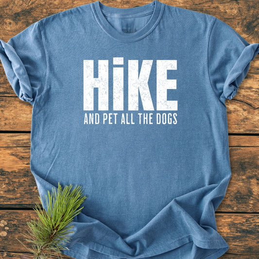 Heather-blue Hike And Pet All The Dogs - Vintage Wash Tee on wood, featuring distressed white lettering. The t-shirt is accompanied by a pine sprig, emphasizing its cozy, relaxed fit and durable make.
