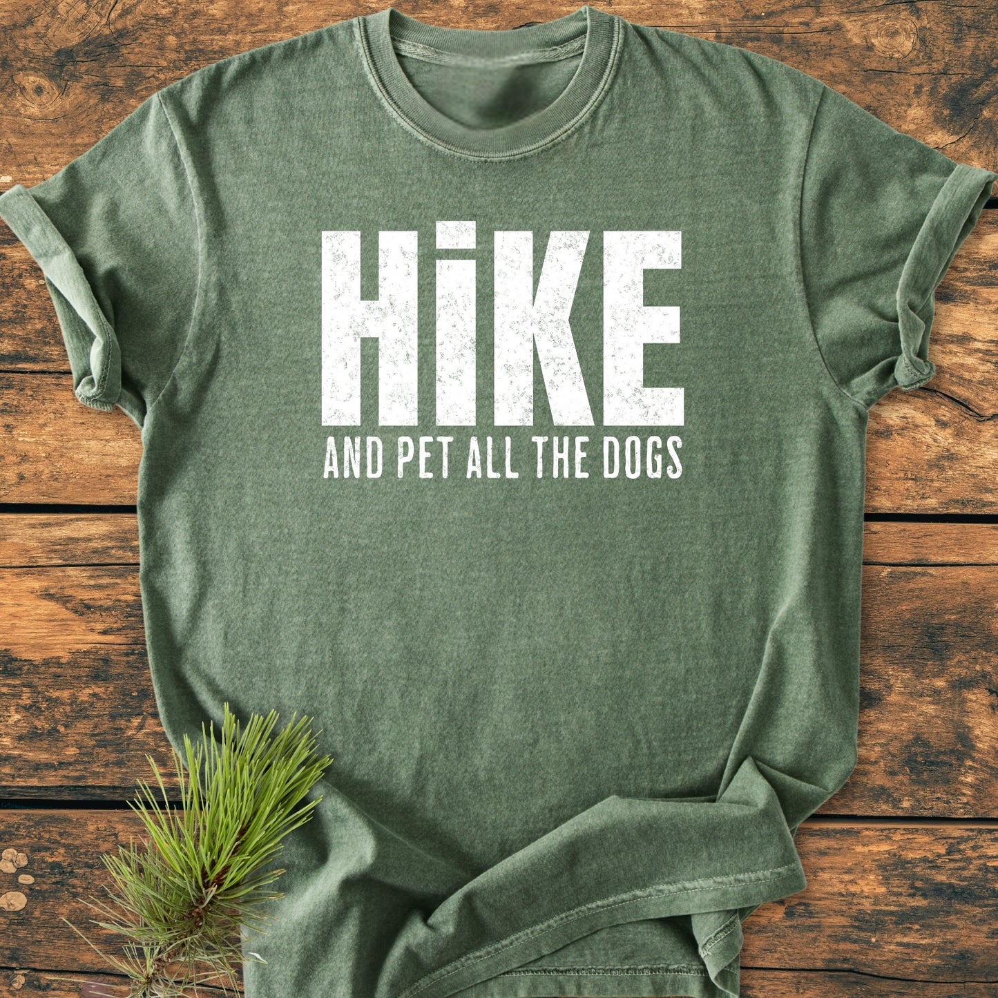 Heather-green Hike And Pet All The Dogs - Vintage Wash Tee with rolled sleeves, displayed on wood. Features distressed white slogan, pine sprig accent, showcasing its rustic, casual charm.