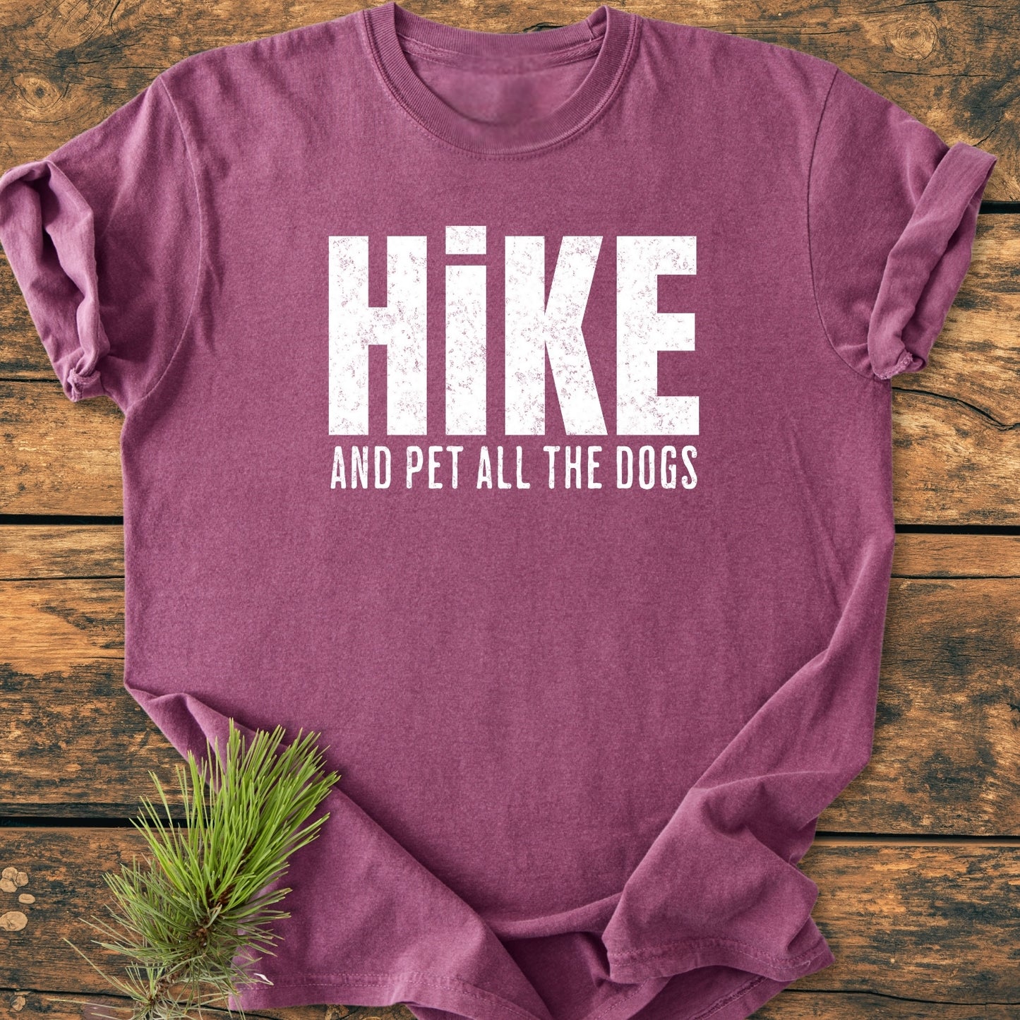 Hike And Pet All The Dogs - Vintage Wash Tee, folded on wood, features distressed white lettering and a pine needle sprig. Soft, relaxed fit, pre-shrunk, ring-spun cotton for comfort and durability.