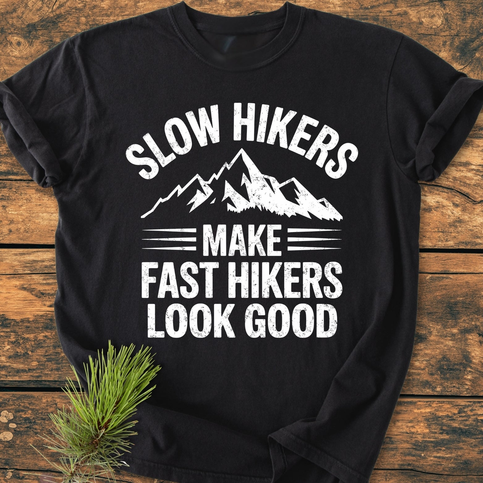 Slow Hikers - Vintage Wash Tee features a white stylized mountain graphic and slogan on a black T-shirt. Laid on a wooden surface with pine sprig, showcasing its cozy, relaxed fit.