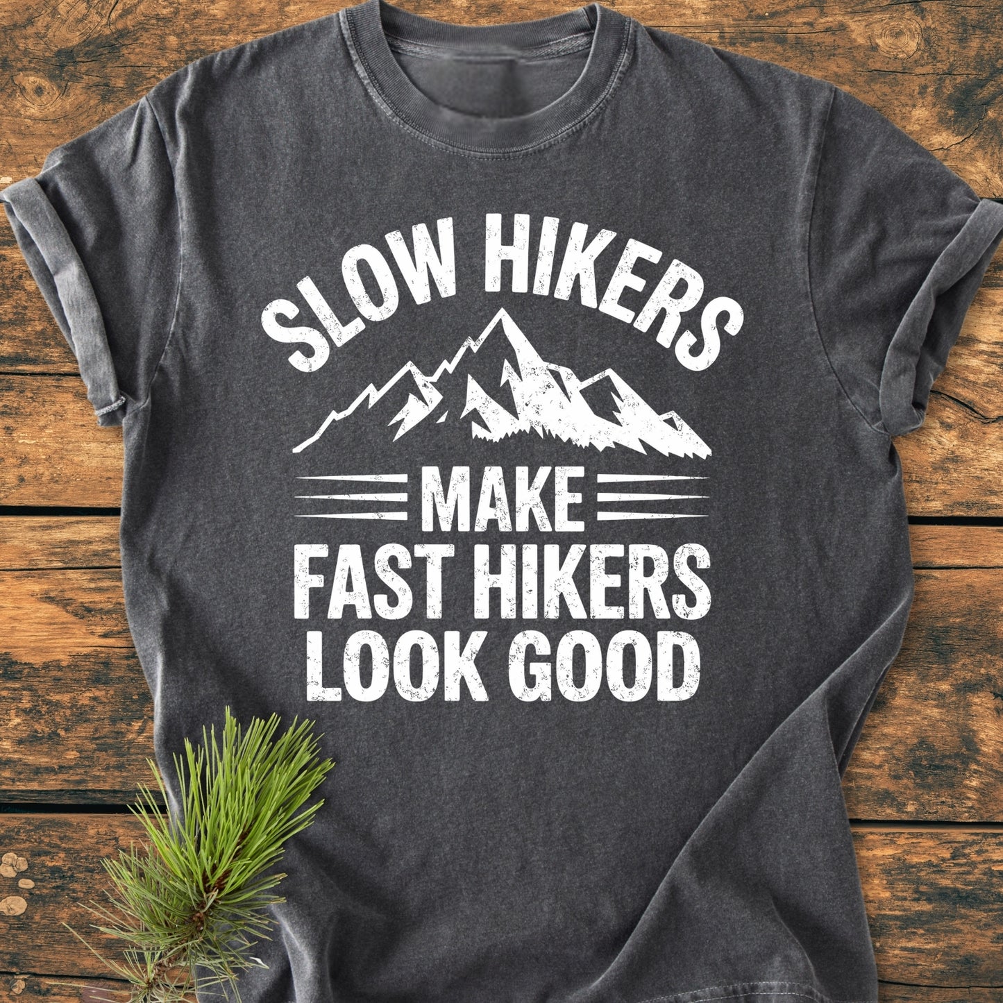 Dark-gray Slow Hikers - Vintage Wash Tee with stylized mountain graphic and slogan, laid on wood. Nearby pine branch accentuates the outdoor theme. Relaxed fit, durable garment-dyed cotton, perfect for daily wear.