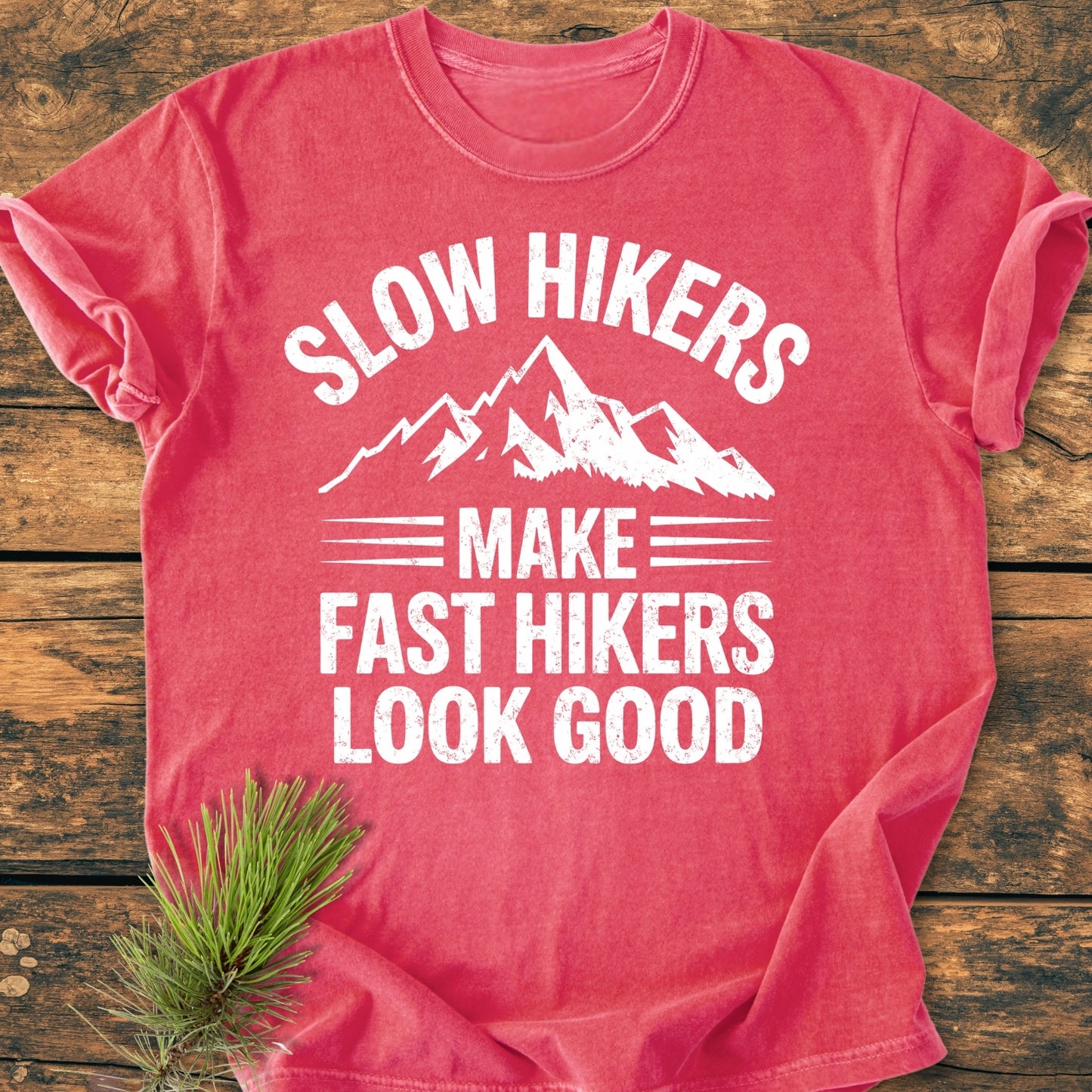 Slow Hikers - Vintage Wash Tee in heather-red, featuring white distressed mountain peaks graphic and playful text, laid flat on wood with pine sprig. Made of 100% ring-spun cotton for comfort.