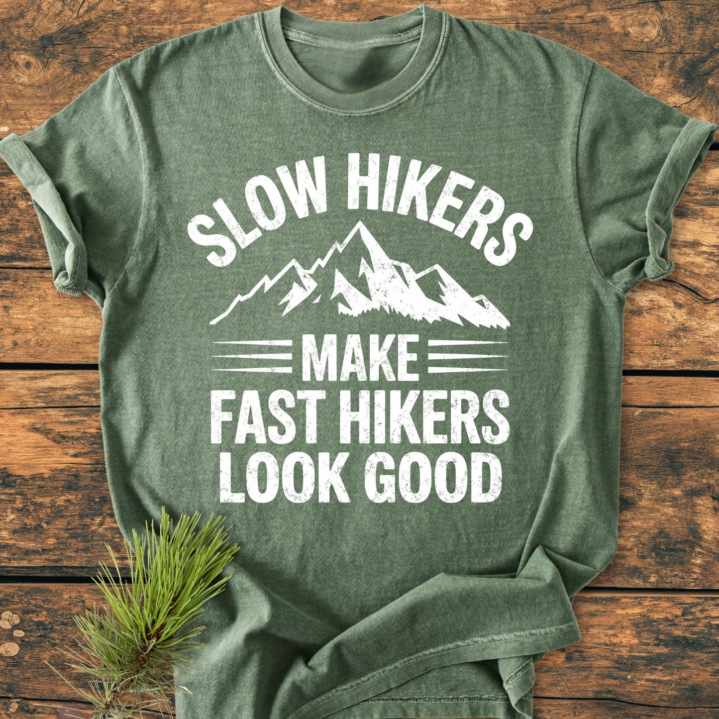 The Slow Hikers - Vintage Wash Tee features a mountain graphic and playful text on a green heathered T-shirt, emphasizing its relaxed fit and outdoor theme, enhanced by a pine sprig accent.