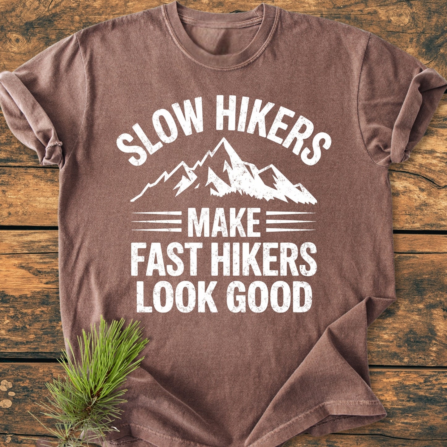 Slow Hikers - Vintage Wash Tee: A brown, short-sleeved shirt with a mountain graphic and slogan, displayed on wood. Features soft, garment-dyed fabric and relaxed fit, ideal for comfortable daily wear.