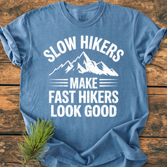 Slow Hikers - Vintage Wash Tee features a white mountain graphic and slogan on a slate-blue background, laid flat on wood. A pine sprig adds a rustic touch.
