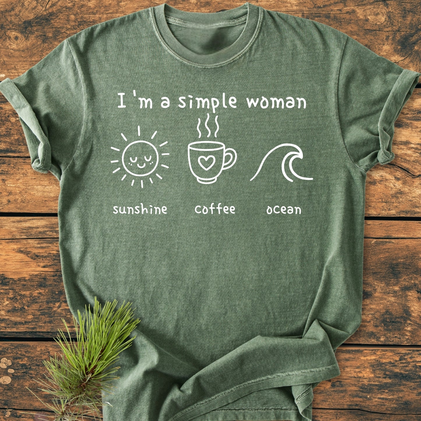 Forest-green I'm A Simple Woman, Sunshine Coffee Ocean - Vintage Wash Tee featuring white screen-print icons of sun, coffee mug, and wave, laid flat on a rustic wooden surface with pine needles.