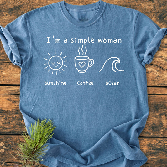 Heather-blue I'm A Simple Woman, Sunshine Coffee Ocean - Vintage Wash Tee with white text and icons of a sun, coffee mug, and ocean wave, displayed flat on a wooden surface.