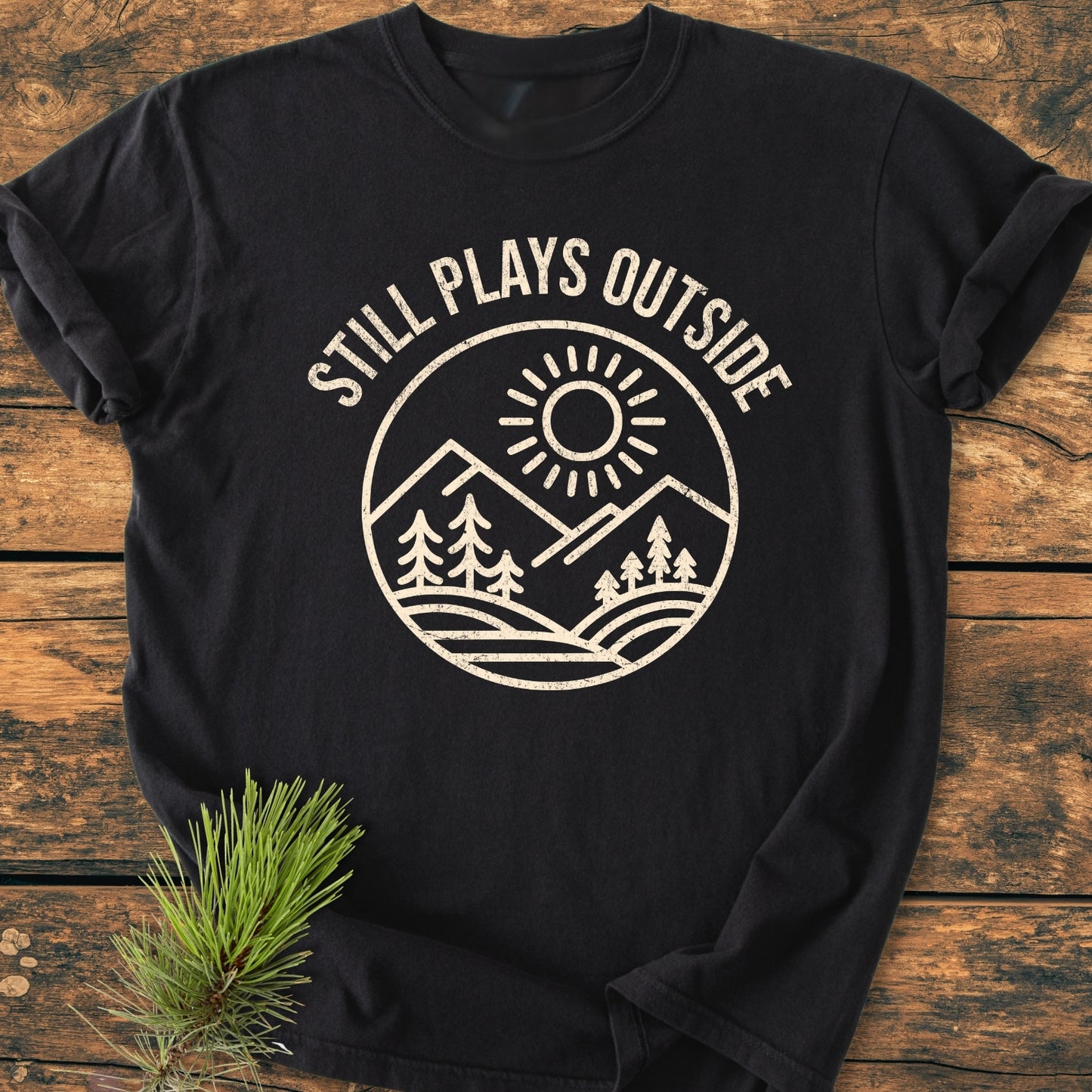 Still Plays Outside - Vintage Wash Tee featuring a circular graphic of mountains and trees. Laid on wood with a pine sprig. Made of 100% ring-spun cotton for comfort and durability.