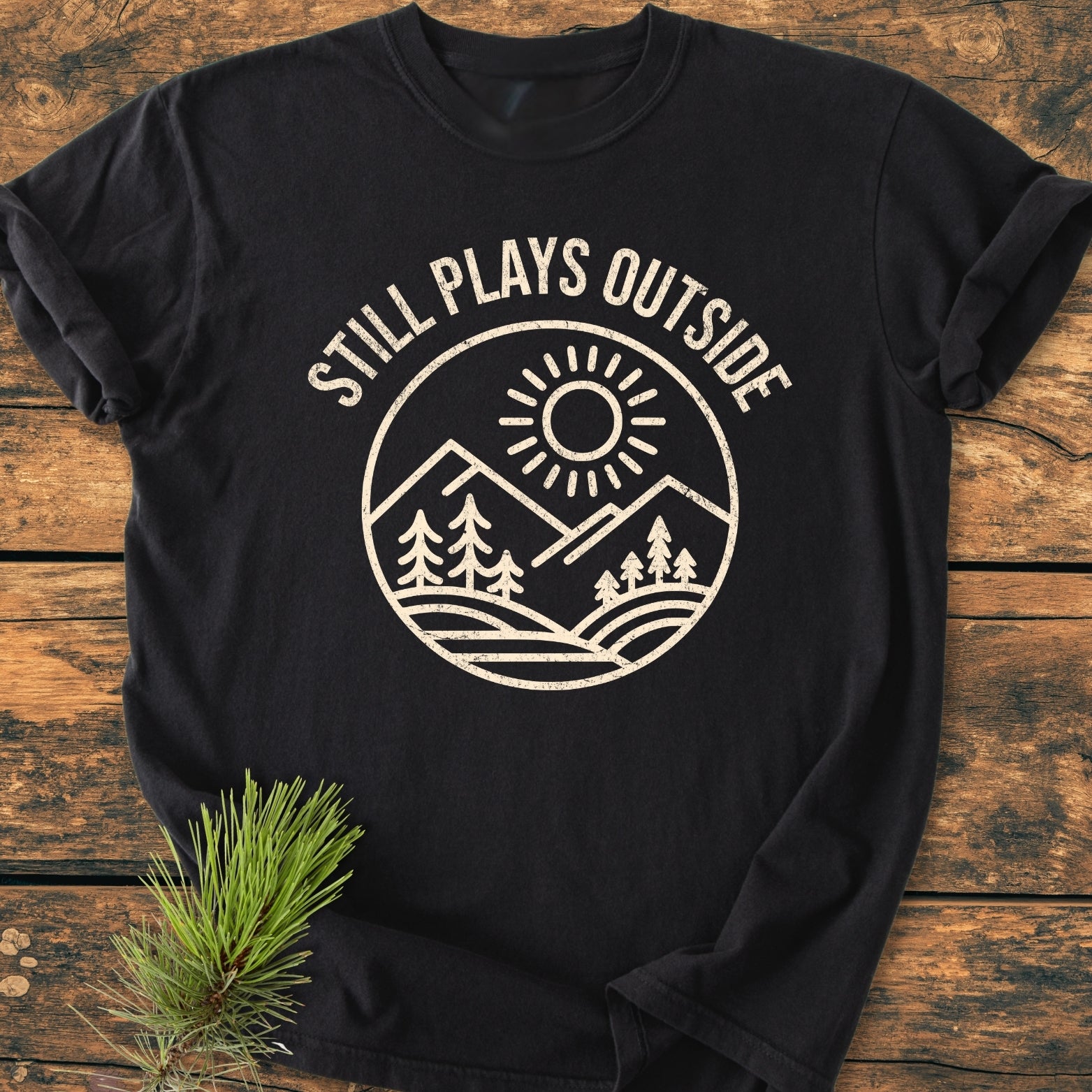Still Plays Outside - Vintage Wash Tee featuring a circular graphic of mountains and trees. Laid on wood with a pine sprig. Made of 100% ring-spun cotton for comfort and durability.