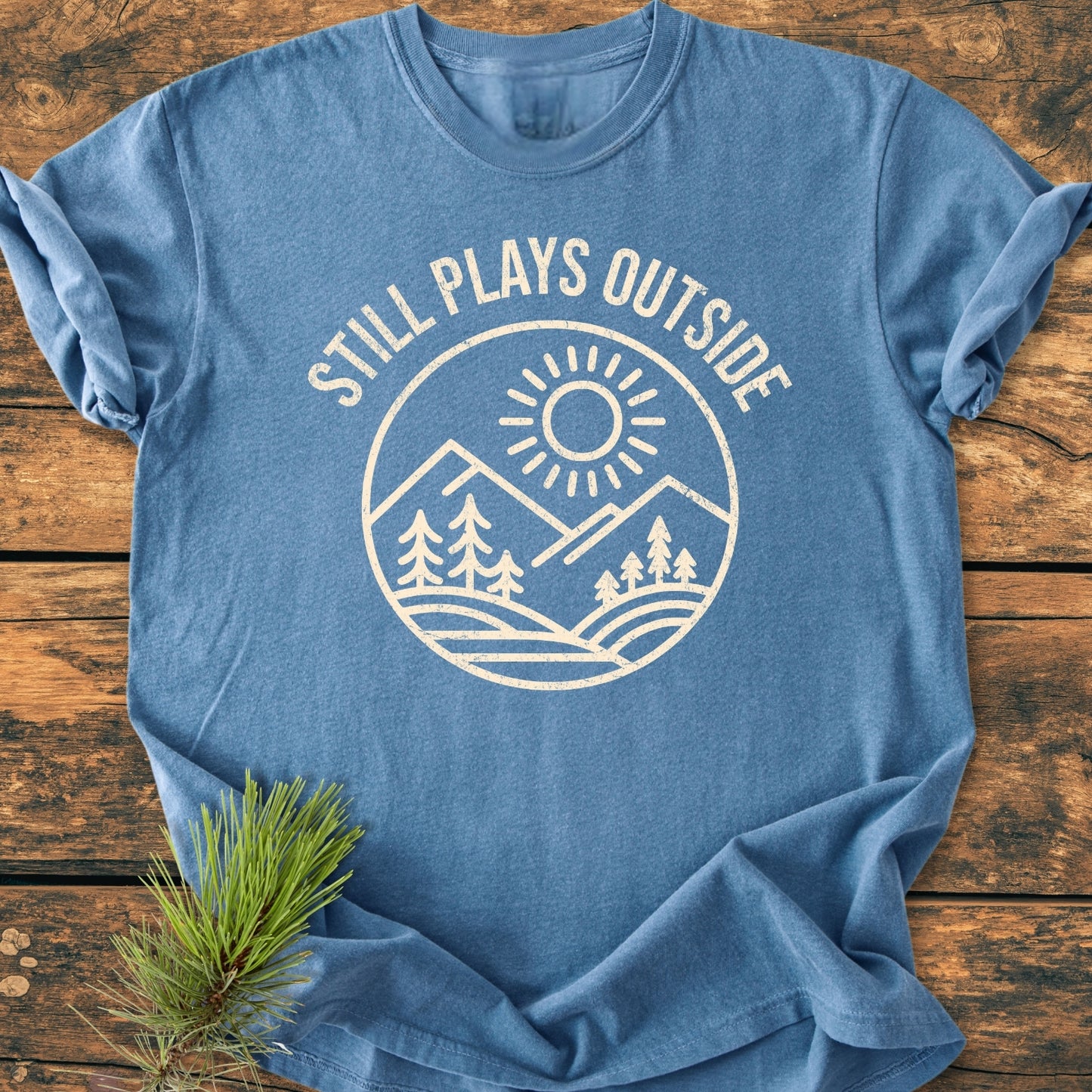 Still Plays Outside - Vintage Wash Tee featuring a circular graphic with mountains and pine trees on a soft, garment-dyed fabric, laid flat on a wood-plank background with a pine sprig nearby.