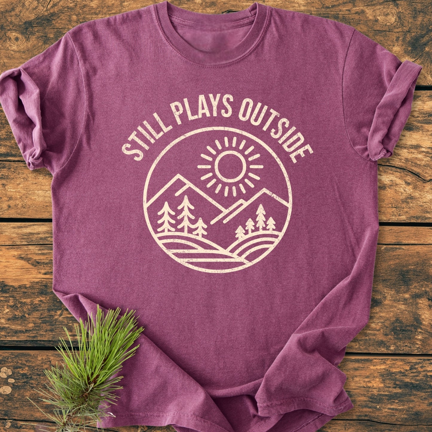 Still Plays Outside - Vintage Wash Tee: A folded purple-heather T-shirt with pine tree and mountain graphic, laid on wood. Features slogan, soft-washed fabric, relaxed fit, and pine needle accent.