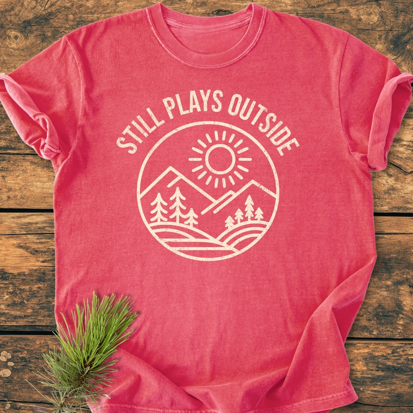 Still Plays Outside - Vintage Wash Tee laid flat on wood, featuring a sun and mountains graphic. The soft, relaxed fit tee is styled with a sprig of evergreen, showcasing its cozy design.