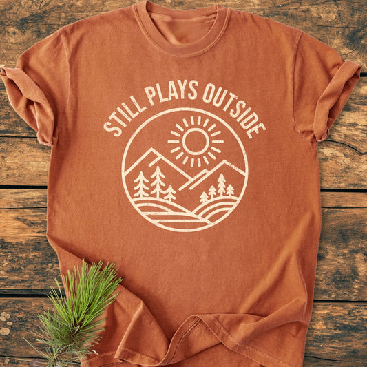 Still Plays Outside - Vintage Wash Tee features a rust color with a sun, mountains, and pine trees graphic. Made of soft, ring-spun cotton, it’s laid on a wooden surface with a pine sprig accent.