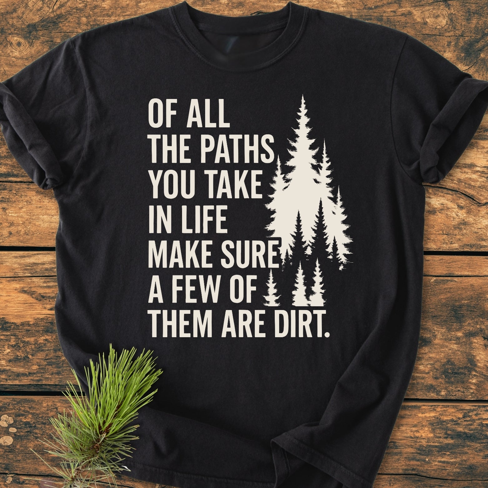 Of All The Paths (John Muir) - Vintage Wash Tee, features a white outdoorsy slogan and pine tree design on a black T-shirt, laid on a wooden surface with a pine sprig beside it.