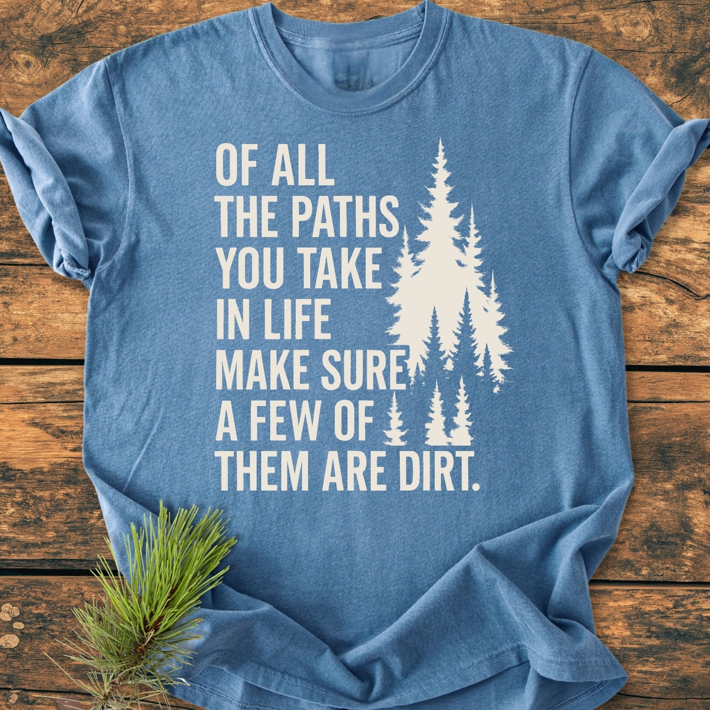 Of All The Paths (John Muir) - Vintage Wash Tee: Heather-blue T-shirt with white slogan and pine-tree silhouettes, laid flat on wood, accented by an evergreen sprig. 100% ring-spun cotton, relaxed fit, durable stitching.