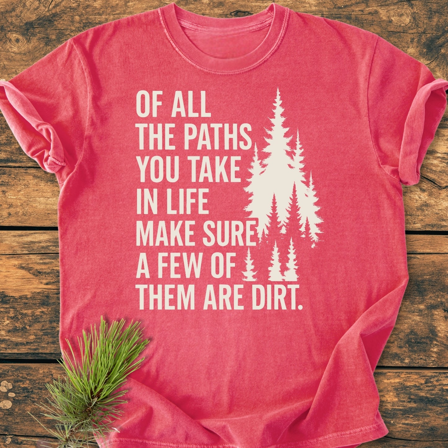 Of All The Paths (John Muir) - Vintage Wash Tee features bold white text and pine silhouettes on a red T-shirt, laid on a wooden surface with a pine needle sprig below.