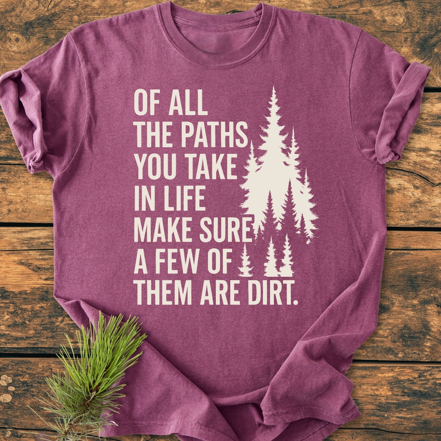 Of All The Paths (John Muir) - Vintage Wash Tee in heather-purple, features an off-white slogan and evergreen tree silhouette, laid on wood with a pine sprig accent, emphasizing its nature theme.